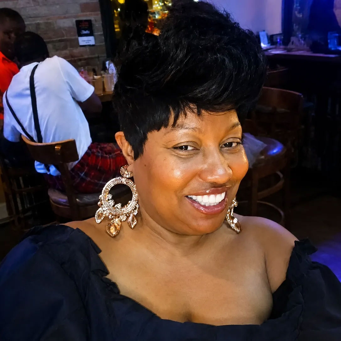 Woman with short black hair, large gold earrings, and a black top smiling at a restaurant or bar.